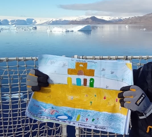 A woman holds up a child's painting on the deck of a ship. In the background is the sea and some icebergs.