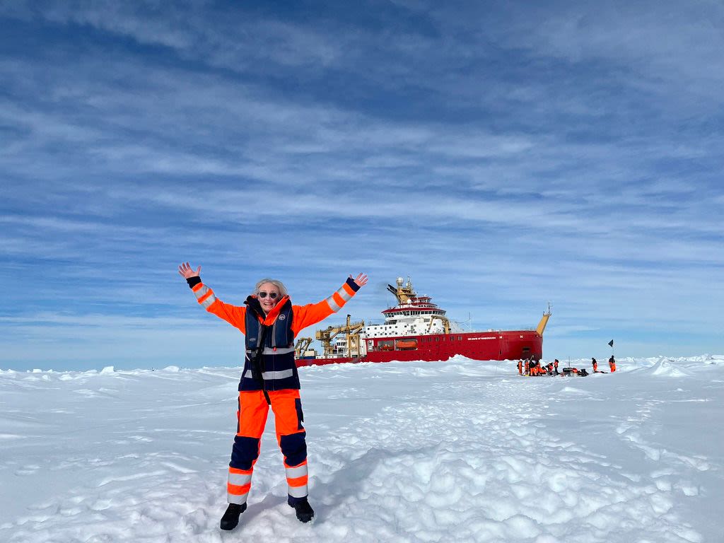 A woman in an orange snowsuit jumps on snowy ground with her hands above her head. In the background there is a red ship and people working on the ice nearby.