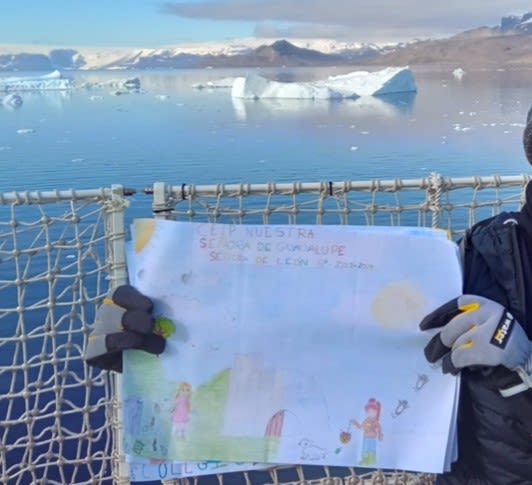 A woman holds up a child's painting on the deck of a ship. In the background is the sea and some icebergs.