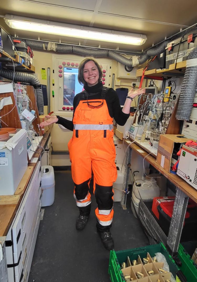A woman in a bright orange work suit, in a lab with lots of scientific equipment.