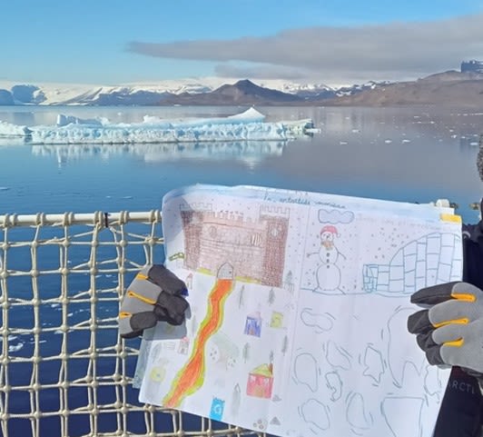 A woman holds up a child's painting on the deck of a ship. In the background is the sea and some icebergs.