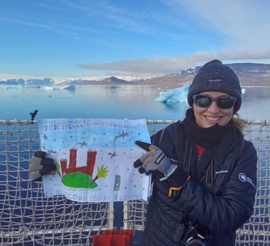 A woman holds up a child's painting on the deck of a ship. In the background is the sea and some icebergs.