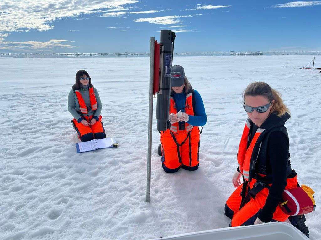 Three women in orange jumpsuits kneel on snowy ground near a pole which has been driven down through ice.