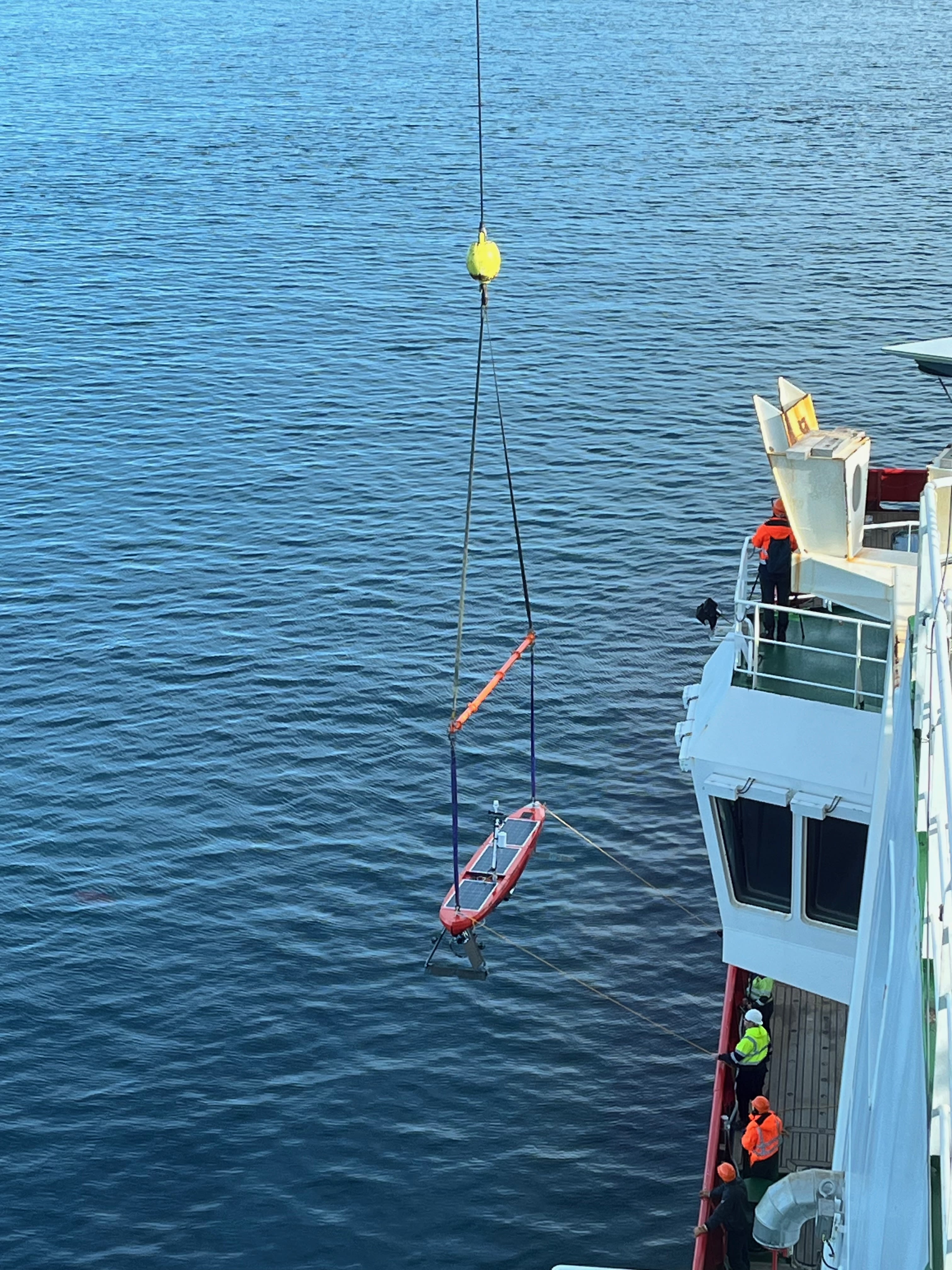 An autonomous surface vessel is lowered by crane into the ocean from a ship.