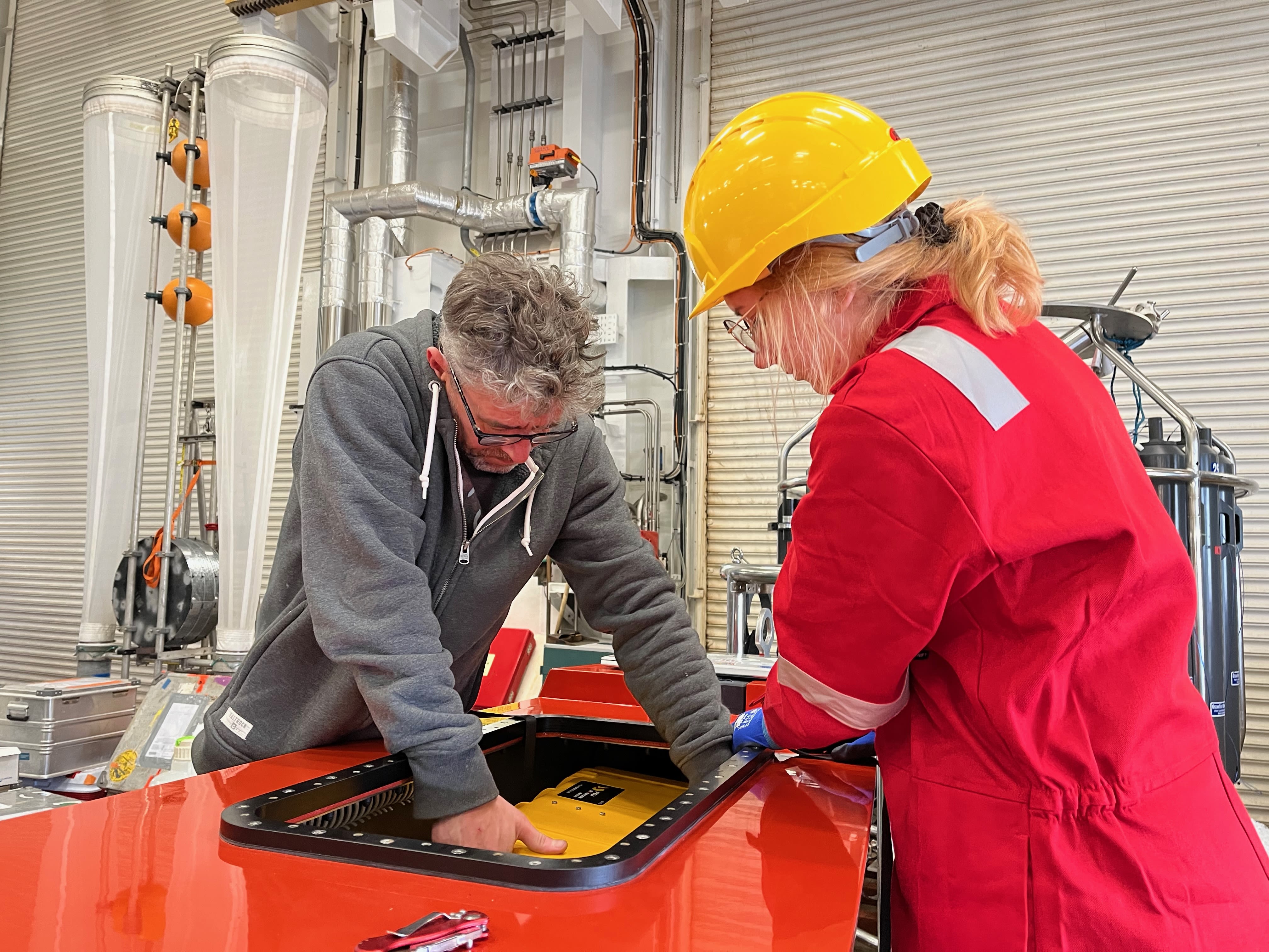 A woman in a red worksuit and a yellow hardhat, and a man in a grey hoodie prepare an autonomous surface vessel.